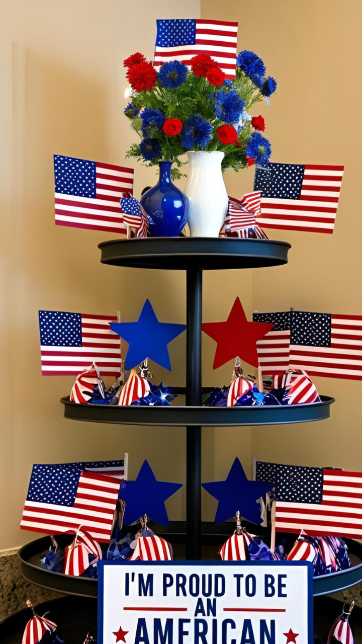 A festive tiered tray decorated with multiple small American flags, red and blue star accents, patriotic-themed bows, and floral arrangements in red, white, and blue. A sign at the bottom reads 'I'm Proud to Be an American.