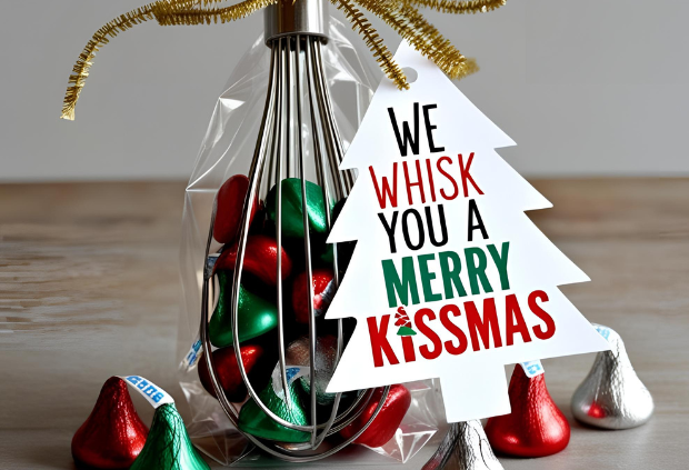 A metal whisk filled with red, green, and silver Hershey's Kisses, wrapped in a clear gift bag with a gold ribbon, and a Christmas tree-shaped tag that reads 'We Whisk You a Merry Kissmas'.